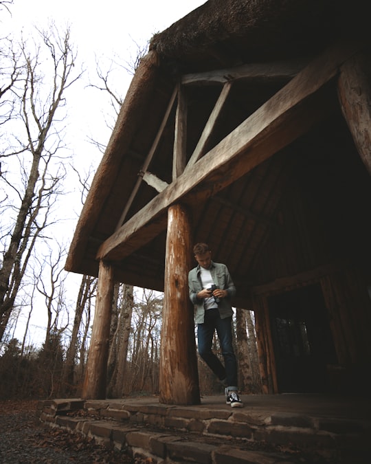 man in white shirt and black pants standing on brown wooden house during daytime in Grimbosq France