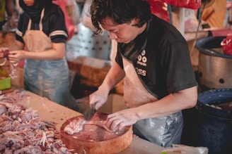 A person is preparing meat on a wooden cutting board in what appears to be a market setting. They are focused on their task, using a large knife to cut through the meat. Another person nearby is also handling meat, wearing a darker uniform with a hair covering. The area is filled with meat and other market items.