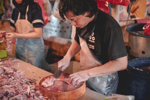 A person is preparing meat on a wooden cutting board in what appears to be a market setting. They are focused on their task, using a large knife to cut through the meat. Another person nearby is also handling meat, wearing a darker uniform with a hair covering. The area is filled with meat and other market items.