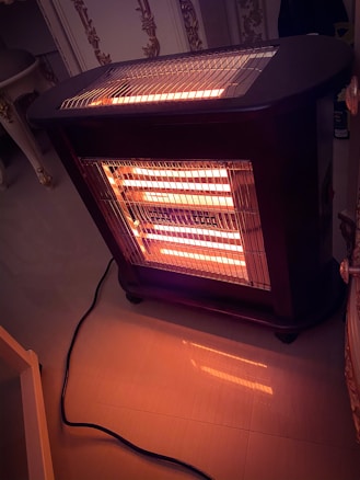 A portable electric heater with visible glowing heating elements is positioned on a polished floor. The heater is plugged in with a black cord extending from it. Ornate furniture and decorative molding can be seen in the background, adding to the cozy indoor atmosphere.