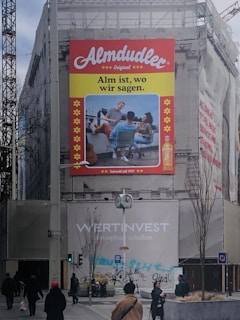 A large advertisement on a building features a bold red 'Almdudler' logo at the top. Below is an image of three people seated outdoors, enjoying a conversation. The slogan 'Alm ist, wo wir sagen.' is displayed prominently. The setting is a construction area with scaffolding and building wraps at street level, where several pedestrians are walking. A clock and street signs are part of the urban landscape.
