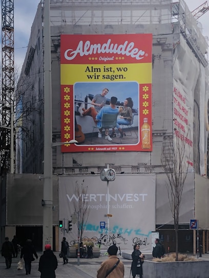 A large advertisement on a building features a bold red 'Almdudler' logo at the top. Below is an image of three people seated outdoors, enjoying a conversation. The slogan 'Alm ist, wo wir sagen.' is displayed prominently. The setting is a construction area with scaffolding and building wraps at street level, where several pedestrians are walking. A clock and street signs are part of the urban landscape.