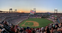 A bustling baseball stadium packed with spectators, illuminated by the warm hues of a setting sun. The field is well-maintained, marked clearly with bases, and surrounded by numerous advertisements. Large screens display game information, while the city skyline is visible in the background.