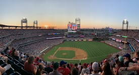 A bustling baseball stadium packed with spectators, illuminated by the warm hues of a setting sun. The field is well-maintained, marked clearly with bases, and surrounded by numerous advertisements. Large screens display game information, while the city skyline is visible in the background.