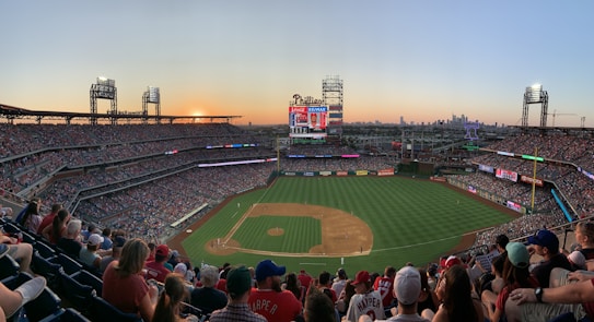 A bustling baseball stadium packed with spectators, illuminated by the warm hues of a setting sun. The field is well-maintained, marked clearly with bases, and surrounded by numerous advertisements. Large screens display game information, while the city skyline is visible in the background.