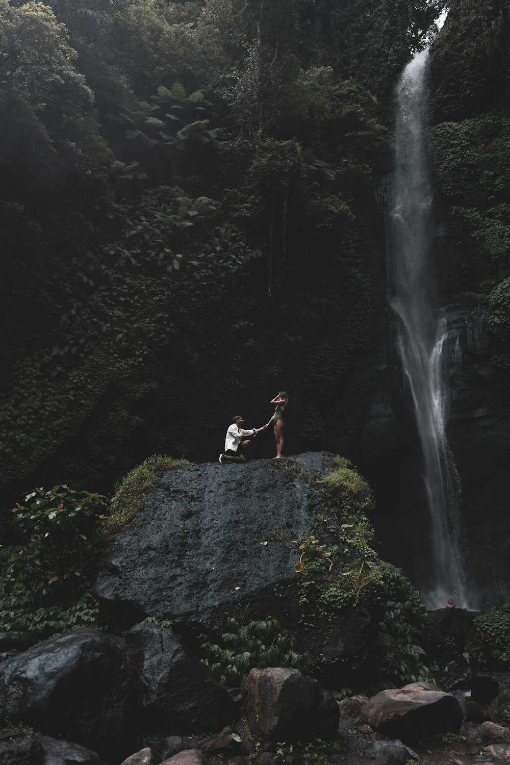 man in white shirt and black shorts standing on rock near waterfalls during daytime