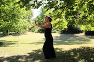 A person in a black outfit appears to be practicing tai chi or a similar movement in a lush, green park. The setting is serene, with sunlight filtering through the leaves of overhanging tree branches, casting dappled shadows on the grass.