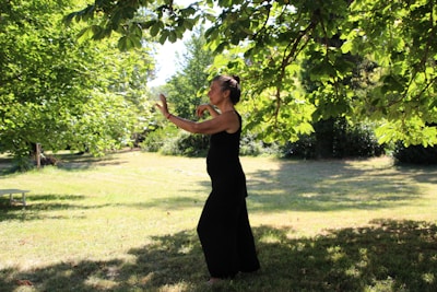 A person in a black outfit appears to be practicing tai chi or a similar movement in a lush, green park. The setting is serene, with sunlight filtering through the leaves of overhanging tree branches, casting dappled shadows on the grass.