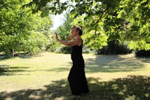 A person in a black outfit appears to be practicing tai chi or a similar movement in a lush, green park. The setting is serene, with sunlight filtering through the leaves of overhanging tree branches, casting dappled shadows on the grass.