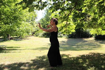 A person in a black outfit appears to be practicing tai chi or a similar movement in a lush, green park. The setting is serene, with sunlight filtering through the leaves of overhanging tree branches, casting dappled shadows on the grass.