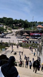 A scene with numerous people walking and interacting in an open area surrounded by various structures and greenery. In the foreground, a person with a black headscarf is descending stairs. The middle ground features a courtyard with several people, some structures with thatched roofs, and a red stage setup with a banner. In the background, there is a forested area with several small buildings.