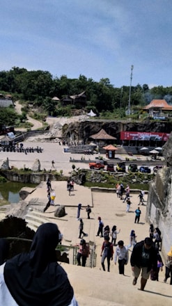 A scene with numerous people walking and interacting in an open area surrounded by various structures and greenery. In the foreground, a person with a black headscarf is descending stairs. The middle ground features a courtyard with several people, some structures with thatched roofs, and a red stage setup with a banner. In the background, there is a forested area with several small buildings.