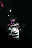 A DJ is performing in a dimly lit club environment, focusing intently on the turntable and mixer. Vibrant lighting effects create an atmospheric glow, bathing the equipment in shades of green and purple.