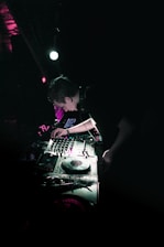 A DJ is performing in a dimly lit club environment, focusing intently on the turntable and mixer. Vibrant lighting effects create an atmospheric glow, bathing the equipment in shades of green and purple.