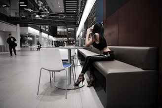 A stylish woman wearing black leather pants and heels sits at a modern, minimalist dining area. The surrounding space is spacious and industrial in design, with large glass panels and a staircase. The background includes a person walking while holding a drink, adding a casual contrast to the otherwise sleek setting.