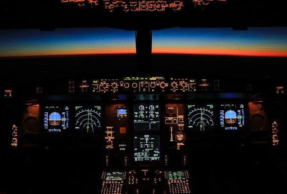 The cockpit of an aircraft is illuminated by soft lights and glowing instrument panels, displaying various screens and gauges. Outside, through the cockpit windows, the horizon is visible with a vibrant gradient from dark blue to a warm orange, suggesting either sunrise or sunset.