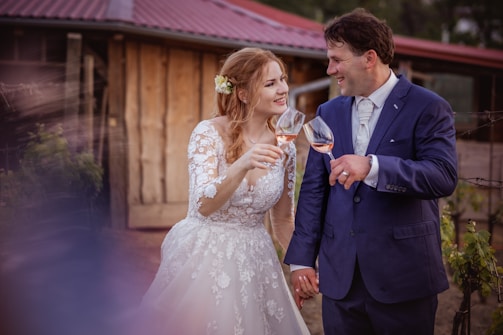A couple dressed in formal attire shares a joyful moment outdoors. The woman wears a white lace wedding dress with floral patterns and a floral hair accessory, while the man dons a navy blue suit with a tie. They are holding wine glasses filled with rosé wine and smiling at each other. The background features a rustic building with a wooden exterior and a red roof.