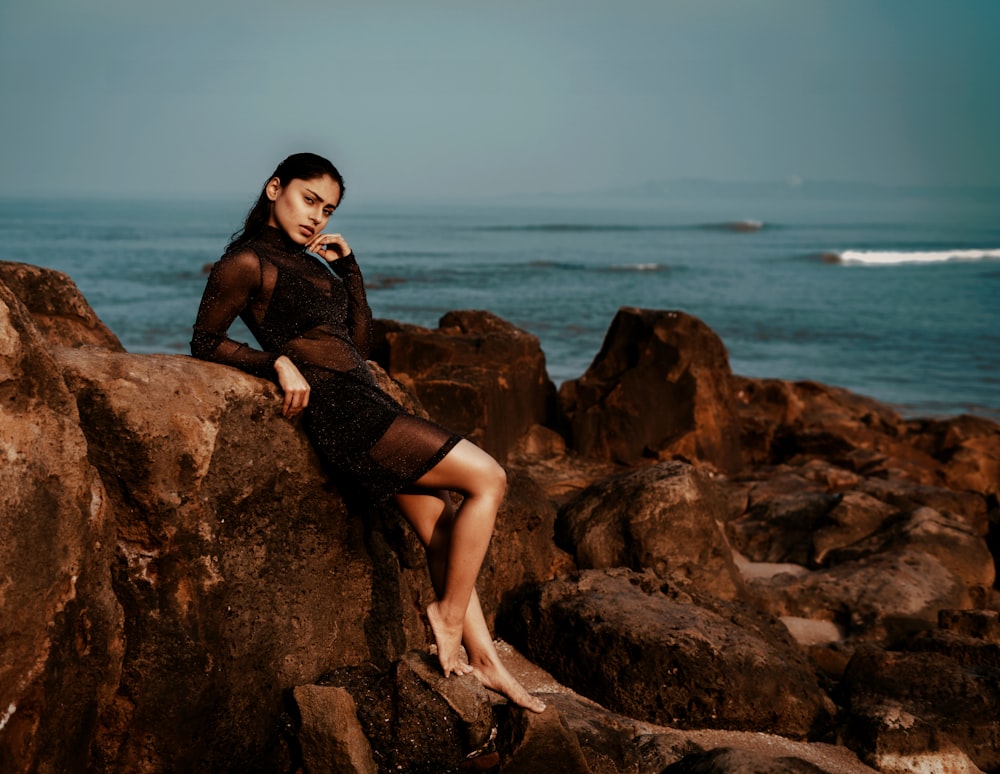 woman in black dress sitting on brown rock near sea during daytime