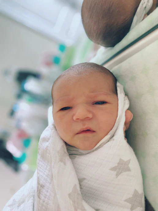A newborn baby is swaddled in a white blanket with gray star patterns, looking intently at the camera. The background is softly blurred, and there is a mirror reflecting part of the baby's head.