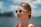 A person wearing white cat-eye sunglasses and large hoop earrings is standing outdoors. The background is blurred, showing hints of blue sky and water. The individual is wearing a red top and has their hair tied back.