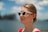 A person wearing white cat-eye sunglasses and large hoop earrings is standing outdoors. The background is blurred, showing hints of blue sky and water. The individual is wearing a red top and has their hair tied back.