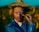 A person with a painted face dressed in a vibrant blue clothing holds a paintbrush near their face. They are wearing a colorful hat that matches their artistic attire. The background features sunflowers and a bright blue sky, setting a vivid and artistic scene.