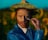A person with a painted face dressed in a vibrant blue clothing holds a paintbrush near their face. They are wearing a colorful hat that matches their artistic attire. The background features sunflowers and a bright blue sky, setting a vivid and artistic scene.