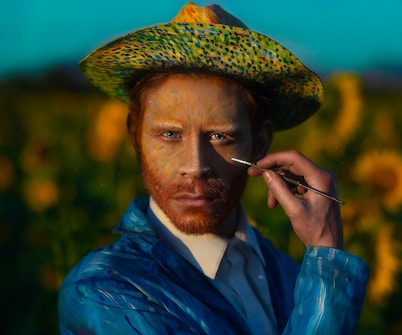 A person with a painted face dressed in a vibrant blue clothing holds a paintbrush near their face. They are wearing a colorful hat that matches their artistic attire. The background features sunflowers and a bright blue sky, setting a vivid and artistic scene.