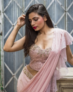 A woman with long hair poses gracefully in a light pink, embellished sari. The background features a geometric metal gate in muted tones, adding a contrasting industrial feel to her elegant attire. Her makeup is subtly glamorous with bright red lips, highlighting her serene expression.