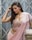 A woman with long hair poses gracefully in a light pink, embellished sari. The background features a geometric metal gate in muted tones, adding a contrasting industrial feel to her elegant attire. Her makeup is subtly glamorous with bright red lips, highlighting her serene expression.