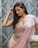 A woman with long hair poses gracefully in a light pink, embellished sari. The background features a geometric metal gate in muted tones, adding a contrasting industrial feel to her elegant attire. Her makeup is subtly glamorous with bright red lips, highlighting her serene expression.