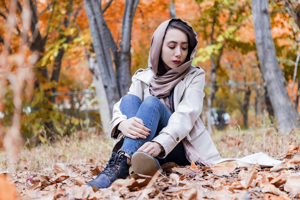 woman in beige coat and blue denim jeans sitting on dried leaves during daytime