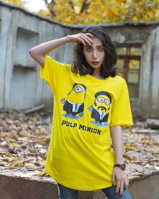 A woman with dark hair is wearing a bright yellow oversized t-shirt featuring cartoon characters styled like minions from a popular animated movie. The shirt reads 'Pulp Minion' and is set against a background of autumn leaves with a rustic structure.