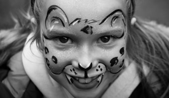 A close-up of a child's face painted with tiger-like patterns in a black and white image. The paint covers the forehead, cheeks, and nose, with intricate designs resembling a cat or tiger face. The expression is joyful and playful.