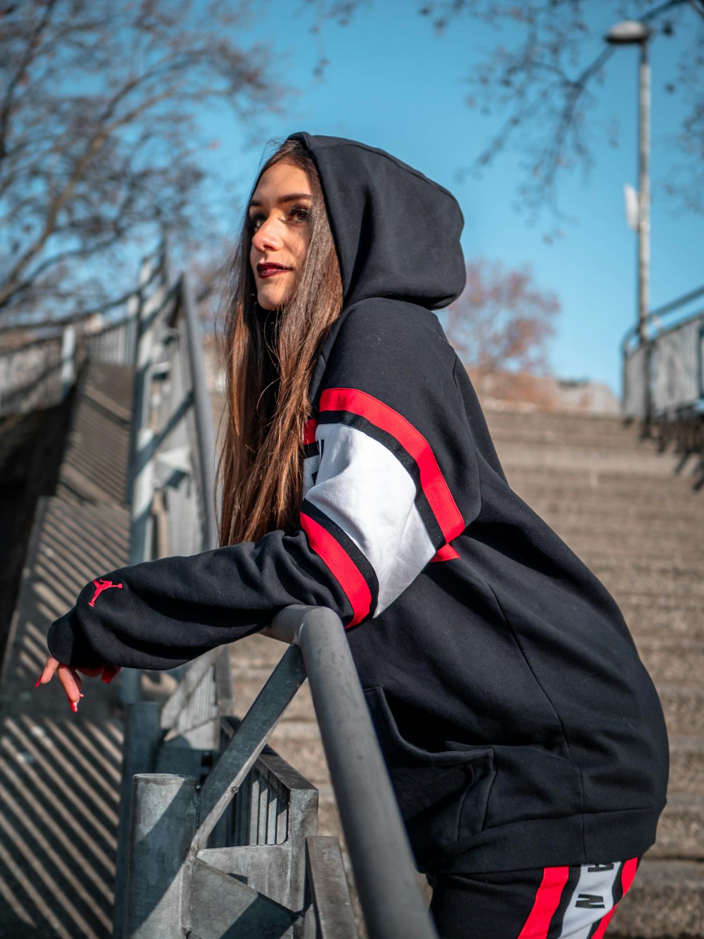 Femme en sweat à capuche noir et rouge debout près d’un arbre nu brun pendant la journée