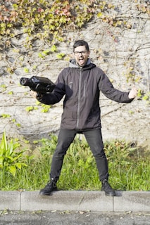 A person with glasses is energetically posing with a large video camera, wearing a black jacket and standing on a curb surrounded by grass and a stone wall with autumn leaves.
