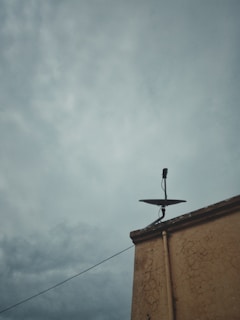 A cloudy sky looms over a building with a satellite dish installed on the edge. The building's wall shows signs of age with visible cracks. The atmosphere seems heavy and overcast, suggestive of an impending storm.