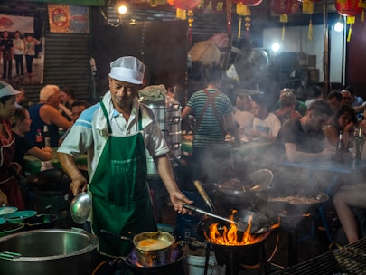 A bustling street food market at night featuring a cook in a green apron and white cap, tending to a flaming stove with several frying pans. Surrounding him are diners seated at tables, enjoying meals. Red lanterns are hanging above, while steam and smoke fill the air, creating an energetic and lively atmosphere.