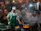 A bustling street food market at night featuring a cook in a green apron and white cap, tending to a flaming stove with several frying pans. Surrounding him are diners seated at tables, enjoying meals. Red lanterns are hanging above, while steam and smoke fill the air, creating an energetic and lively atmosphere.