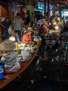 A bustling indoor market scene with vendors on wooden boats selling various goods like fruits, vegetables, and packaged items. The vendors wear traditional hats and are illuminated by soft hanging lights, while people walk around browsing and buying items.