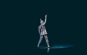 A person dressed in a colorful, striped suit stands on a dark stage. They are facing away and have one arm raised in a dramatic pose, with light illuminating their figure.