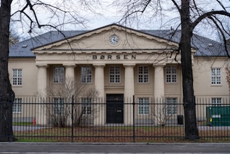 A large, classical building with a Greek-style facade and four prominent columns at the entrance. The word 'BØRSEN' is displayed prominently at the top of the building. Surrounding the structure is a black wrought-iron fence and leafless trees, with sparse vegetation at the front.