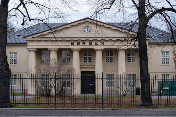 A large, classical building with a Greek-style facade and four prominent columns at the entrance. The word 'BØRSEN' is displayed prominently at the top of the building. Surrounding the structure is a black wrought-iron fence and leafless trees, with sparse vegetation at the front.