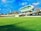 A cricket stadium featuring a large, modern, and futuristic-looking observation deck or media center suspended above the stands. The grass field is vast and well-maintained, and the sky is blue with some clouds. Trees and a tall structure, possibly a chimney or tower, are visible in the background.