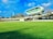 A cricket stadium featuring a large, modern, and futuristic-looking observation deck or media center suspended above the stands. The grass field is vast and well-maintained, and the sky is blue with some clouds. Trees and a tall structure, possibly a chimney or tower, are visible in the background.