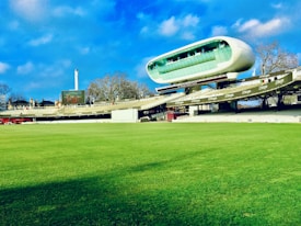 A cricket stadium featuring a large, modern, and futuristic-looking observation deck or media center suspended above the stands. The grass field is vast and well-maintained, and the sky is blue with some clouds. Trees and a tall structure, possibly a chimney or tower, are visible in the background.