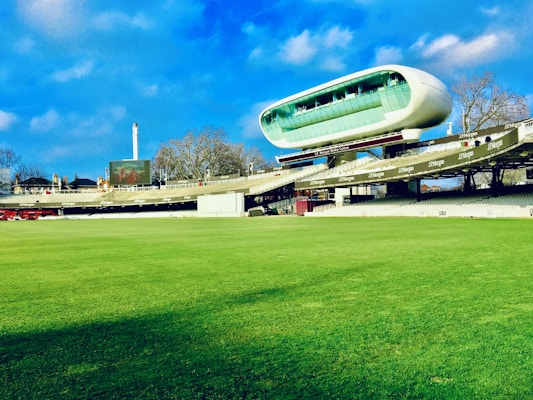 A cricket stadium featuring a large, modern, and futuristic-looking observation deck or media center suspended above the stands. The grass field is vast and well-maintained, and the sky is blue with some clouds. Trees and a tall structure, possibly a chimney or tower, are visible in the background.