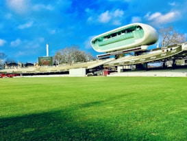 A cricket stadium featuring a large, modern, and futuristic-looking observation deck or media center suspended above the stands. The grass field is vast and well-maintained, and the sky is blue with some clouds. Trees and a tall structure, possibly a chimney or tower, are visible in the background.