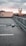 A drone hovers in focus above an empty rooftop parking lot with a blurred cityscape in the background under a cloudy sky.