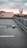 A drone hovers in focus above an empty rooftop parking lot with a blurred cityscape in the background under a cloudy sky.